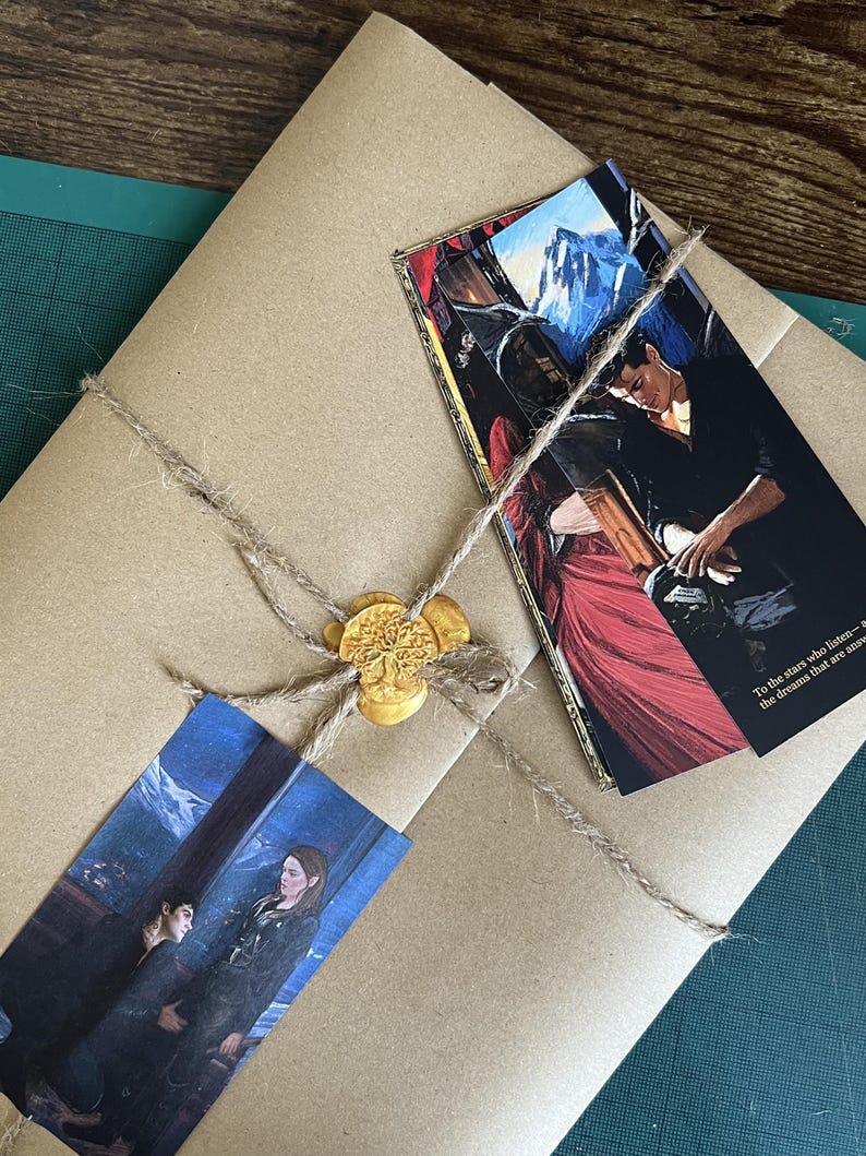 May include: A brown paper package tied with twine and sealed with a gold wax seal featuring a tree design. Two rectangular bookmarks are tucked under the twine, each displaying a colorful illustration of two figures and text.