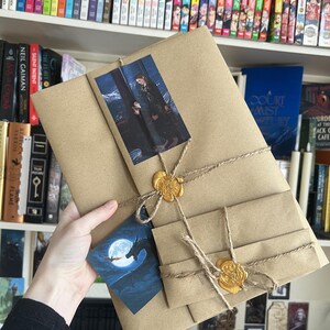 May include: A rectangular brown paper package wrapped with twine and secured with two gold wax seals. The package features a rectangular print of two figures and a separate triangular card depicting a dragon silhouetted against a full moon.