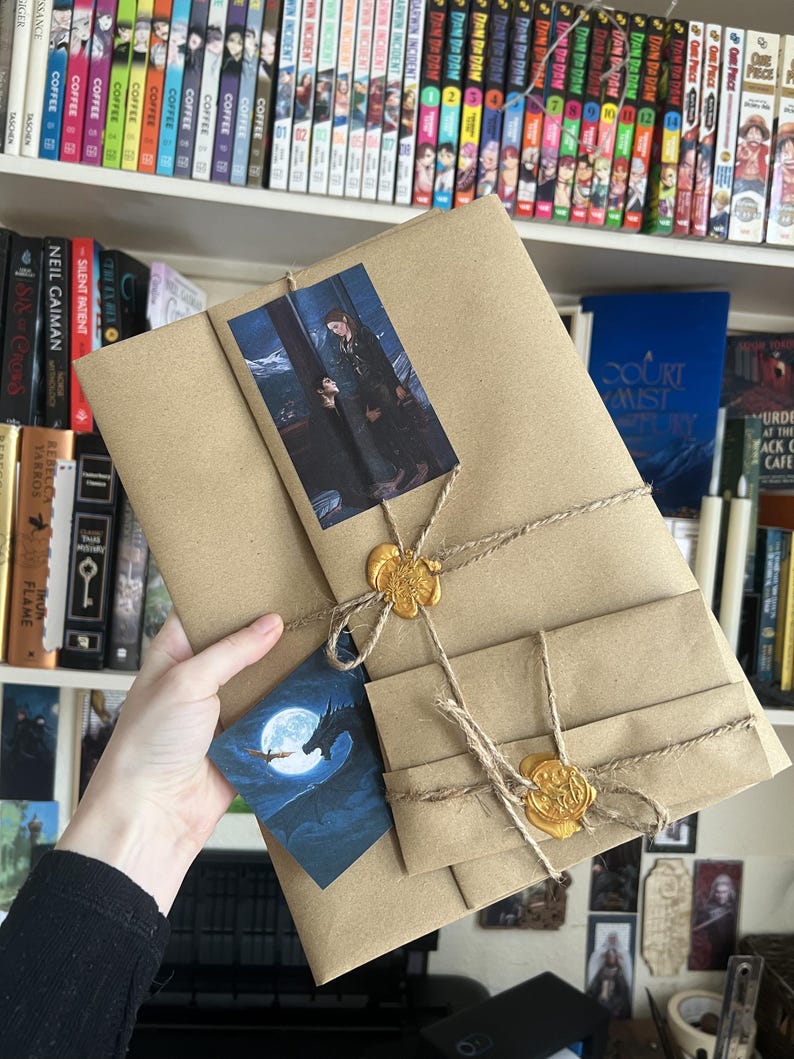May include: A rectangular brown paper package wrapped with twine and secured with two gold wax seals. The package features a rectangular print of two figures and a separate triangular card depicting a dragon silhouetted against a full moon.