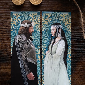 May include: Two bookmarks featuring illustrations of a king and queen. The king is wearing a crown and armor, while the queen is wearing a white gown and a crown. The background is a teal blue with gold floral patterns.
