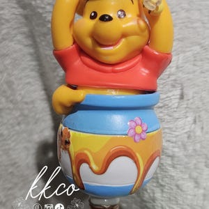 May include: A Winnie the Pooh-themed figurine featuring the character in a red shirt and blue hat, with a honey pot design. The figurine has a bee on its head and a honey-themed charm at the bottom.