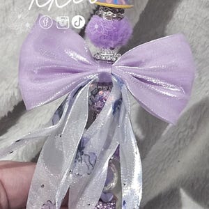 May include: A decorative pen featuring a Chip and Mrs. Potts design. The pen has a purple, rhinestone-covered barrel, a lavender bow, and white ribbons with floral accents. The pen's design includes the text "kkco" and social media icons.