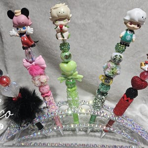 May include: A collection of decorative pens featuring character figurines, beads, and embellishments. The pens have various designs, including Mickey and Minnie Mouse, and a chef. The pens are arranged in a rhinestone holder.