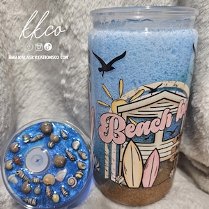 May include: A clear glass jar with a beach-themed design. The jar features a blue and white background with a beach hut, surfboards, and the words "Beach Life". A smaller, round container with seashells is next to the jar. The text "kkco" and website address are visible.