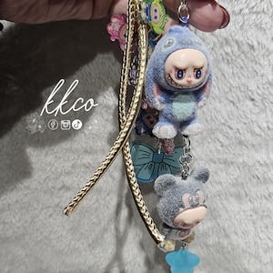 May include: A keychain with multiple charms, including a blue plush figure in a bunny suit, a gray bear, and a green alien. The keychain has a gold-colored cord and a blue chain. The text "kkco" is visible.