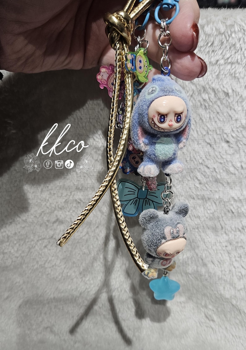 May include: A keychain with multiple charms, including a blue plush figure in a bunny suit, a gray bear, and a green alien. The keychain has a gold-colored cord and a blue chain. The text "kkco" is visible.