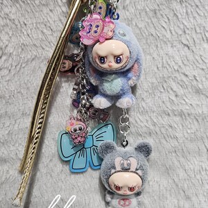 May include: A keychain featuring two plush figures, one resembling a bunny and the other a bear, both with doll-like faces. The keychain includes a blue star charm and a light blue bow-shaped charm. The text "lkco" is visible.