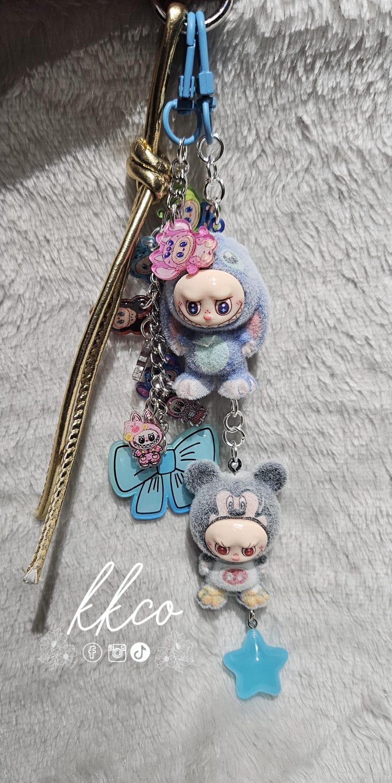May include: A keychain featuring two plush figures, one resembling a bunny and the other a bear, both with doll-like faces. The keychain includes a blue star charm and a light blue bow-shaped charm. The text "lkco" is visible.