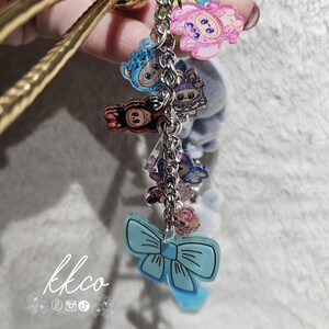 May include: A gold-colored bangle bracelet with a chain of colorful acrylic charms. The charms include cartoon characters and a light blue bow. The text "kkco" is visible in the lower left corner.