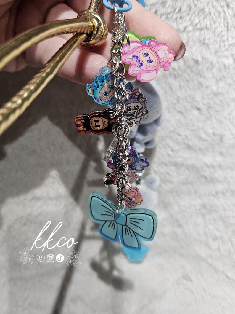 May include: A gold-colored bangle bracelet with a chain of colorful acrylic charms. The charms include cartoon characters and a light blue bow. The text "kkco" is visible in the lower left corner.