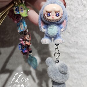 May include: A blue plush keychain featuring a cartoon character with a smiling face and bunny ears. The keychain has a silver chain and a gray plush bear charm. Other colorful charms are attached to a gold cord. The text "kkco" is visible.