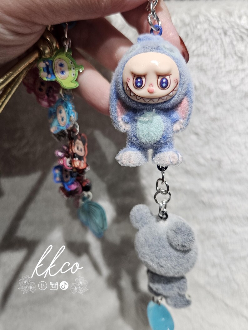 May include: A blue plush keychain featuring a cartoon character with a smiling face and bunny ears. The keychain has a silver chain and a gray plush bear charm. Other colorful charms are attached to a gold cord. The text "kkco" is visible.