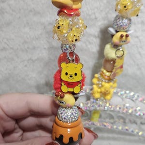 May include: A decorative pen featuring Winnie the Pooh characters. The pen is adorned with yellow, red, and silver embellishments, including a Pooh bear figurine, honey pot, and sparkling beads. The pen has a glittery finish.