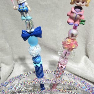 May include: Two decorative pens with doll-like figures and beaded accents. One pen features a blue and white color scheme with a doll holding a red balloon. The other pen has a pink and pearl theme.