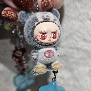 May include: A gray plush keychain featuring a cartoon character with red eyes and a menacing grin. The keychain has a blue star charm and is attached to a beaded chain. The text "kkco" is visible.