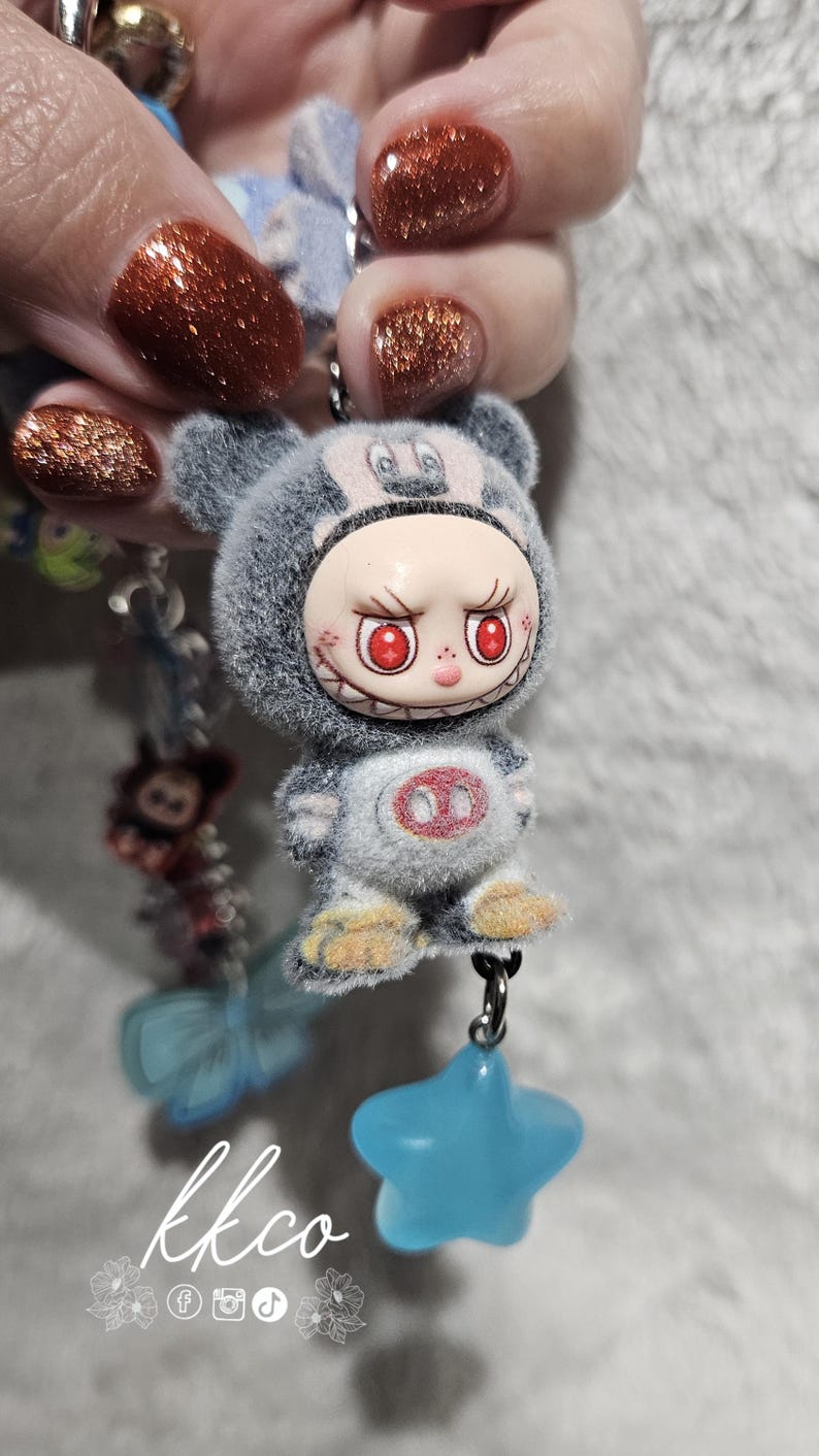 May include: A gray plush keychain featuring a cartoon character with red eyes and a menacing grin. The keychain has a blue star charm and is attached to a beaded chain. The text "kkco" is visible.