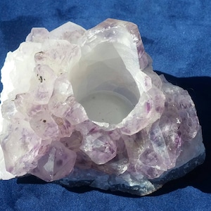 May include: A purple amethyst crystal candle holder with a natural, rough texture. The holder has a hollow center for a candle.