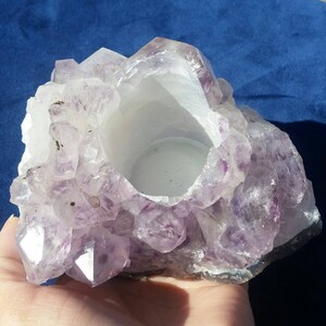 May include: A purple amethyst crystal candle holder with a smooth, white interior. The holder is made of natural amethyst crystals and has a rough, textured exterior.