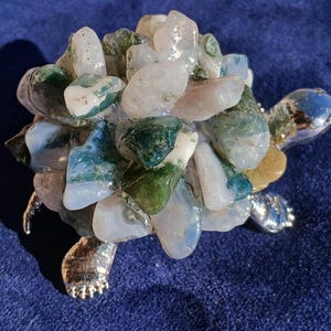 May include: A decorative turtle figurine featuring a shell constructed from numerous polished, tumbled stones in shades of white, translucent gray, and deep forest green. The head, legs, and tail are crafted from a smooth, reflective silver-toned metal.