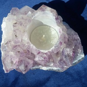 May include: A purple amethyst crystal candle holder with a white tea light candle inside. The holder is made of natural amethyst crystals and has a rough, textured surface.