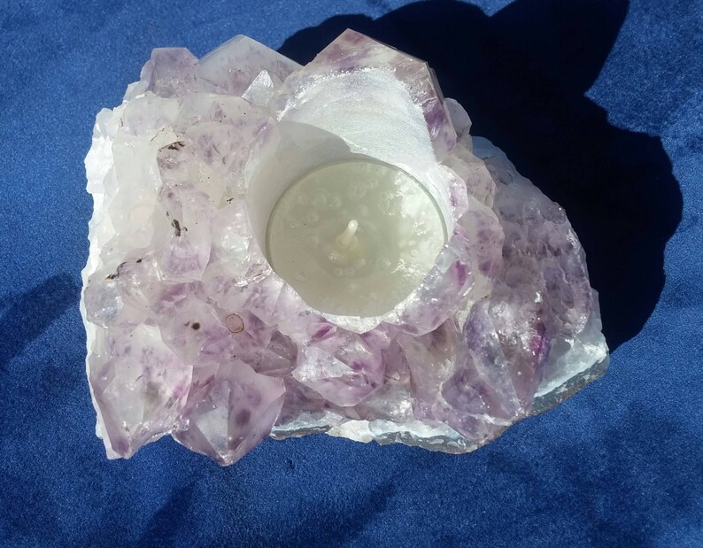 May include: A purple amethyst crystal candle holder with a white tea light candle inside. The holder is made of natural amethyst crystals and has a rough, textured surface.