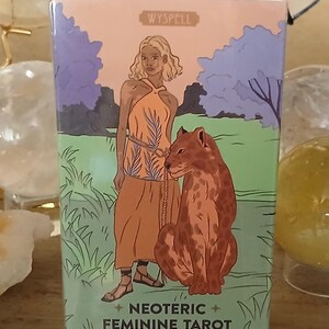 May include: A rectangular tarot deck box titled WYSPELL NEOTERIC FEMININE TAROT tarot deck of 78 cards & guidebook. The front features an illustration of a person in an orange dress standing with a large leopard in a green landscape with purple trees.