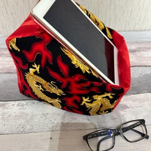 Könnte beinhalten: Ein roter und schwarzer Tablet-Ständer aus Stoff mit einem goldenen Drachenmuster. Der Ständer ist mit einem Tablet darauf aufgestellt und ein Paar schwarze Brillen liegen auf einer Holzoberfläche.