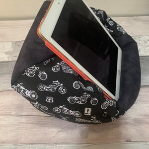 May include: A soft, wedge-shaped tablet stand featuring a black fabric cover with a white motorcycle pattern, a Route 66 sign, and an arrow icon labeled ONLY. The stand supports a white tablet with a thin orange border, resting at an angled position.