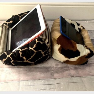 May include: Two tablet stands. One has a giraffe print in black, brown, and tan, the other a cowhide pattern in brown, white, and tan. The stands are designed to hold tablets. The stands are made of fabric.