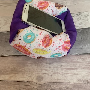 May include: A purple fabric phone holder with a white and colorful donut pattern. A white Samsung smartphone is inside the holder. The holder is made of soft fabric and is designed to keep the phone secure.