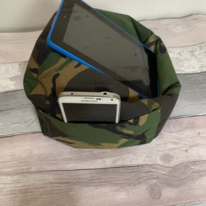 May include: A camouflage fabric pillow with a built-in stand for holding a tablet or phone. The pillow is filled with soft stuffing and has a pocket for storing a phone. The tablet is a black and blue Samsung tablet.