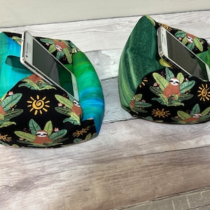 May include: Two green and blue fabric phone stands with a sloth and sun pattern. The stands are filled with stuffing and are designed to hold a phone upright.