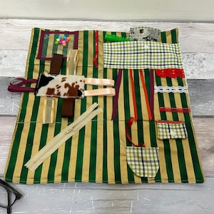 May include: A green and gold striped fabric play mat with various textures and colours. It features a zipper, ribbons, a cow print patch, a pocket, and a clear plastic window with colourful buttons.