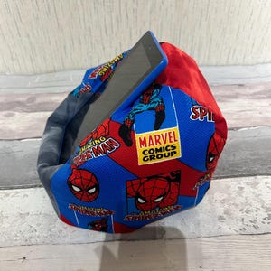 Puede incluir: Un cojín de frijoles azul y rojo con un patrón de Spiderman. El cojín tiene las palabras "The Amazing Spiderman" y "Marvel Comics Group" impresas en él. Una tableta está descansando sobre el cojín.