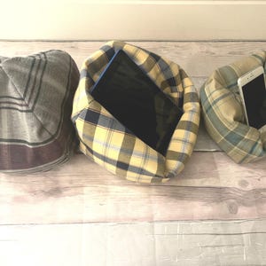 May include: Three fabric pillow-like holders for tablets and phones. The holders are in a plaid pattern. One is grey and white, one is yellow and blue, and one is yellow and green. The yellow and blue holder has a tablet inside, and the yellow and green holder has a phone inside.