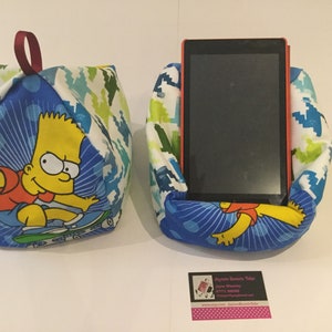 May include: Two fabric tablet holders. One is empty, the other holds a tablet. Both are made of blue fabric with a Bart Simpson graphic. The fabric also has a white and green pattern. The empty holder has a maroon loop.