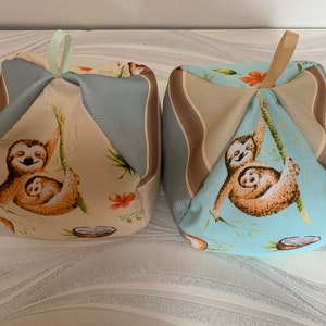 May include: Two beige and blue fabric cubes with a sloth design. Each cube has a loop handle on top. The sloths are brown and have a light blue background. The fabric has a striped pattern.