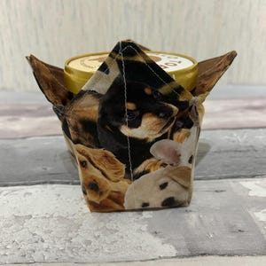 Op de afbeelding: Een stoffen ijsbekerhouder met een patroon van verschillende hondenrassen en -kleuren. De houder is ontworpen om een pint-grootte ijscontainer te bevatten, met bruine flappen die zich vanaf de bovenkant uitstrekken.