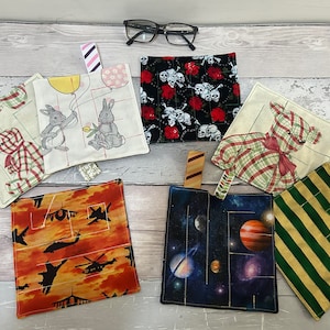 Könnte beinhalten: Sechs verschiedene Stoffquadrate mit verschiedenen Designs, darunter ein Teddybär, Hasen, Totenköpfe und Rosen, ein karierter Hund, eine Weltraumszene und ein grün-gelb gestreiftes Muster. Jedes Quadrat hat eine Schlaufe zum Aufhängen.