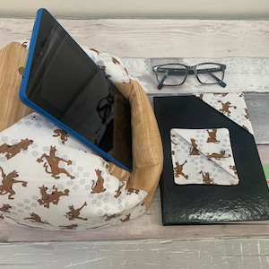 May include: A blue tablet stand with a white fabric cover featuring a cartoon dog print. The stand is holding a black tablet. A black notebook with a white fabric bookmark featuring the same cartoon dog print is next to the stand. A pair of glasses and a small card with text are also visible.