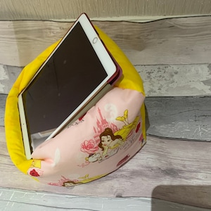 May include: A yellow and pink tablet pillow with a tablet inside. The pillow is made of two fabrics: a solid yellow and a pink fabric with a princess design. The tablet is white with a black screen.