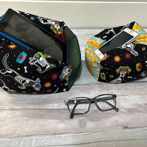 May include: Two fabric phone and tablet stands with a Day of the Dead dog design. The stands are black with colourful flowers and bones. One stand is holding a phone and the other is holding a tablet.