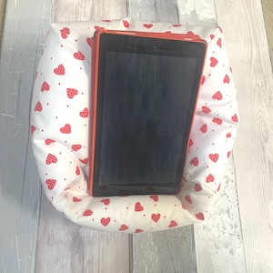 May include: A white pillow with red hearts printed on it. The pillow has a built-in stand for a tablet or phone. The tablet is black and has a red frame.