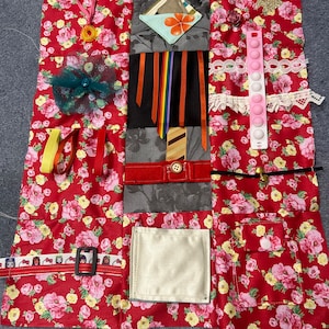 May include: A sensory activity board with diverse textures and elements. The board features red floral fabric, ribbons, a pop-it toy, a buckle, and other tactile components. It is designed to stimulate sensory exploration and fine motor skills.