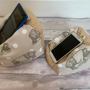 May include: Two beige and brown fabric phone and tablet holders with white polka dots and a brown bear print. The holders are filled with stuffing and have a pocket to hold a phone or tablet.