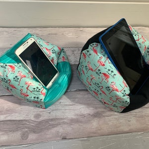 May include: Two fabric phone and tablet holders. One holds a white smartphone, the other a black tablet. Both are made of turquoise fabric with a flamingo and palm tree pattern, and a solid turquoise fabric. The tablet holder has a black base.