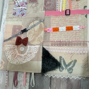 Puede incluir: Una alfombra de actividad de tela beige con varias texturas y elementos para el juego sensorial. Cuenta con una mariposa, un parche de piel, un borde de encaje, un lazo, un collar, una cremallera, un botón, un juguete pop-it, cintas y un bolsillo pequeño.