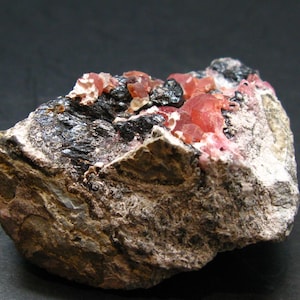 May include: A close-up of a rough mineral specimen. The rock is a mix of brown, grey, and black, with vibrant red and pink crystalline formations. The texture is rugged and uneven, showcasing the raw beauty of the stone.