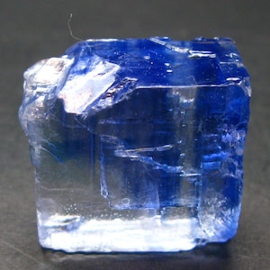 May include: A translucent, blue-hued crystal with a cuboid shape. The crystal's facets reflect light, creating a shimmering effect. The color transitions from clear to deep blue, showcasing its natural beauty. A small, thin fiber is visible on the top.