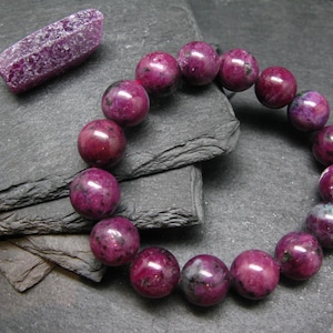 May include: A bracelet of large, round, deep magenta beads with a speckled appearance. A raw, elongated, purple crystal sits nearby. The jewelry rests on a dark gray, textured surface, creating a striking contrast.